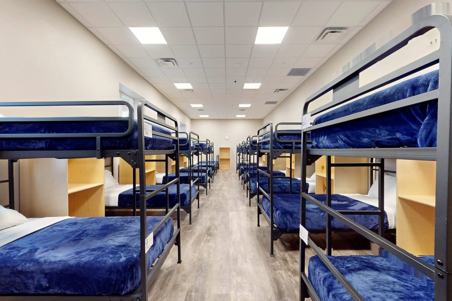 A long shelter sleeping room lined with metal bunk beds made up with navy blankets and wooden storage cubbies between them.