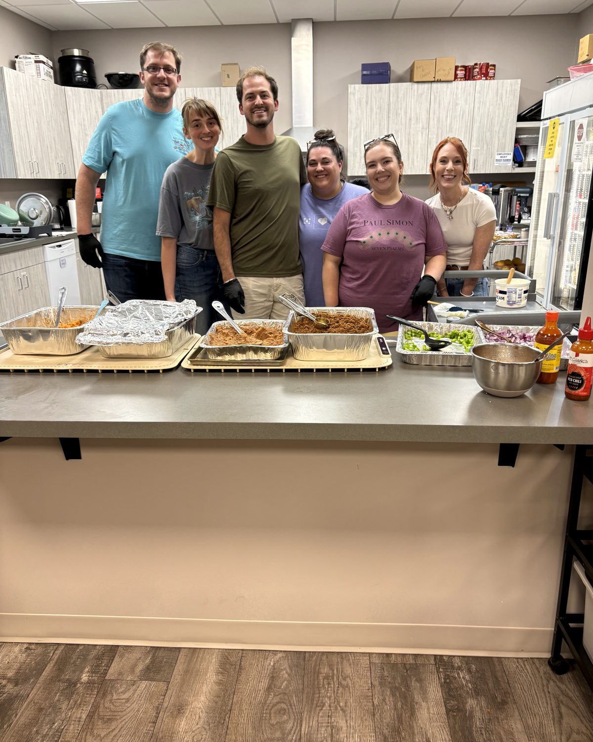Six volunteers from a local group standing behind a counter of foil trays they cooked and brought in for dinner service.