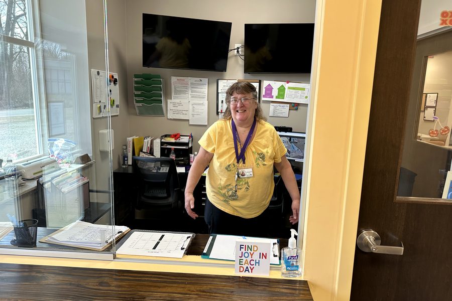 A staff member smiling behind the day center's front desk window, with sign-in sheets on the counter and a note reading "find joy in each day."