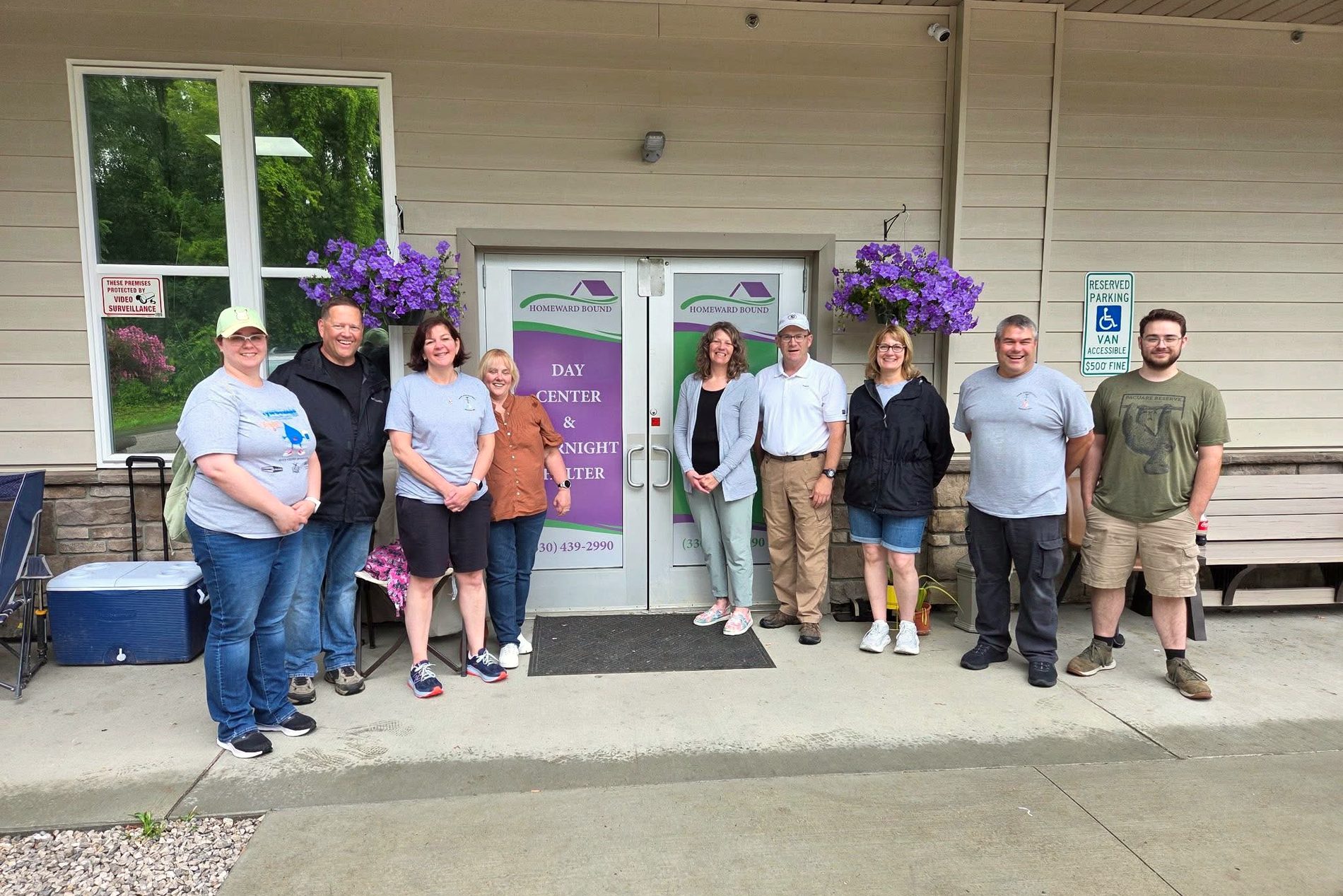 Nine Homeward Bound staff and volunteers standing outside the front doors of the day center and overnight shelter, with hanging baskets of purple flowers on either side.