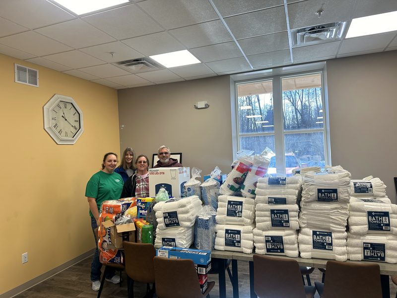 Volunteers standing behind tables stacked with donated toilet paper, paper towels, and household supplies from a drive.