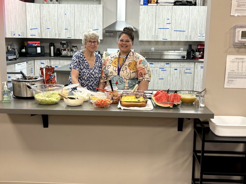 Two volunteers behind the kitchen counter with bowls of salad, fruit, and watermelon laid out for a meal.