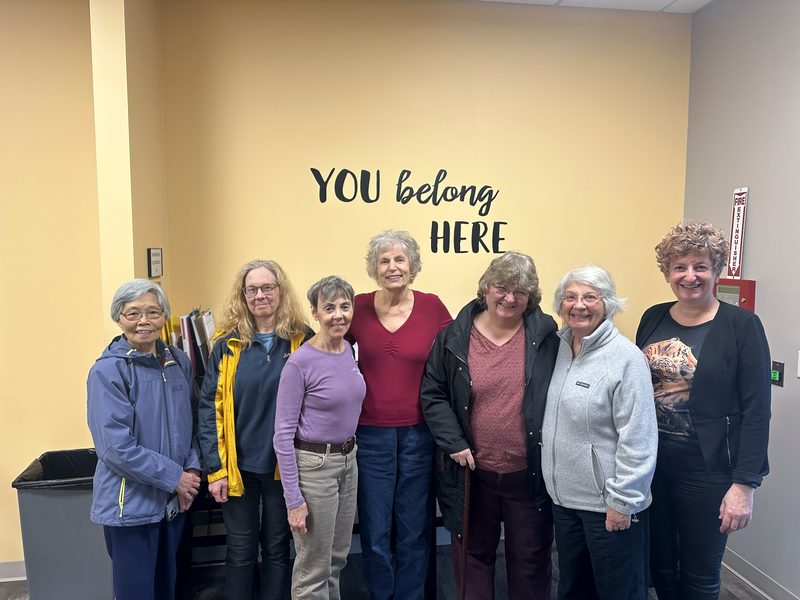 Seven women standing together in front of the painted "You belong here" wall inside the day center.