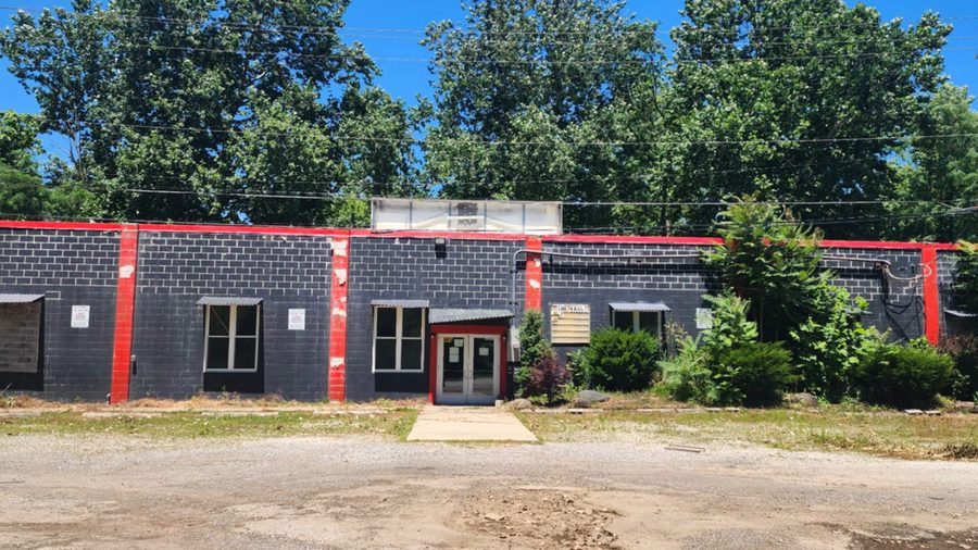 The vacant building at 981 Grosjean Road before renovation: painted grey block walls with red pillars, boarded windows, and overgrown shrubs.