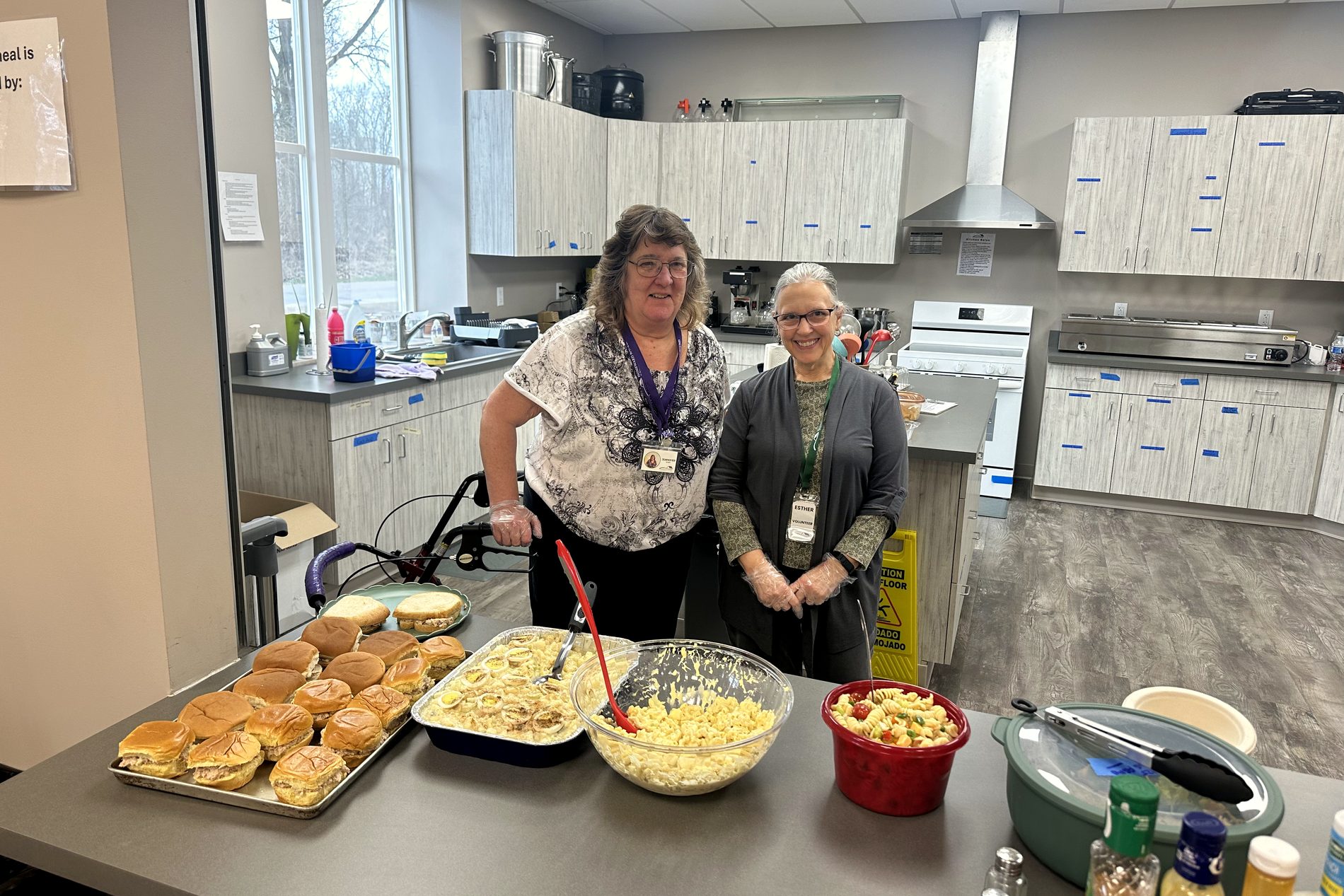 Two Homeward Bound staff members standing behind a counter of sandwiches, casserole, and sides plated up for the evening meal.