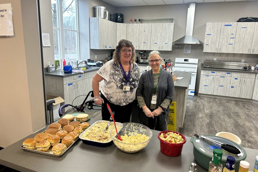 Two volunteers standing behind a kitchen counter laid out with trays of sandwiches, casserole, and salad ready to serve.