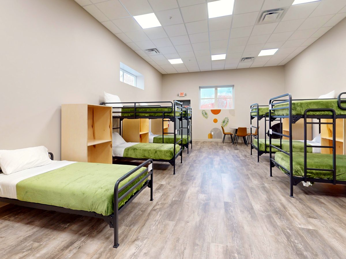 A bright shelter sleeping room at Homeward Bound with made-up single and bunk beds, wooden storage cubbies, and a window at the far end.