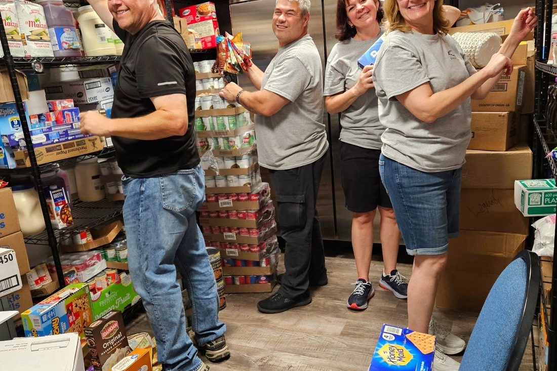 Four volunteers restocking the pantry shelves with tinned food, cereal, and drinks, turning to smile at the camera.